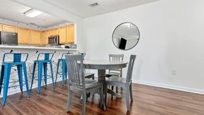 Cocina moderna con armarios de madera, microondas y refrigerador. Hay un comedor con una mesa redonda y sillas azules.