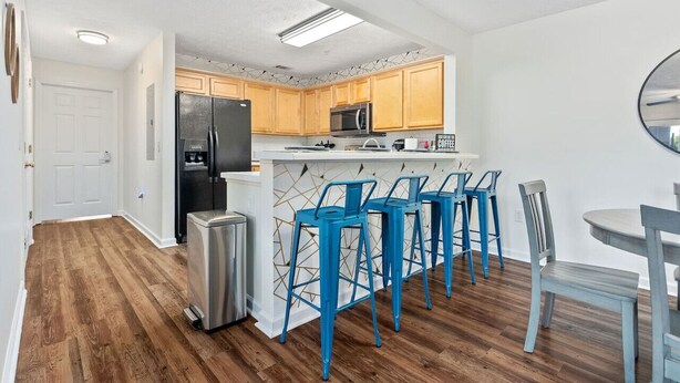 Una cocina moderna con armarios de madera, un refrigerador negro, un microondas y un lavamanos de acero inoxidable. Hay taburetes altos azules y una pequeña mesa con sillas.