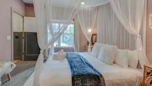 Een slaapkamer met een groot bed, witte gordijnen en een raam met uitzicht op groen.