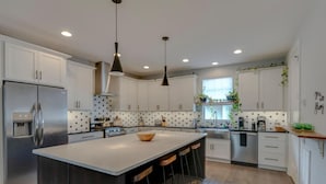 Een moderne keuken met witte kasten, een roestvrijstalen koelkast, een groot eiland met krukken, hanglampen en een spatwand met stippen.
