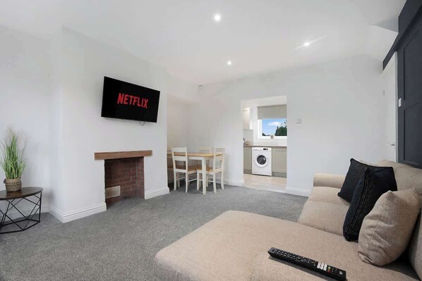 A modern living room with a flat-screen TV, a sofa, a fireplace, a dining area, and a kitchen visible through an open door. - 3BR in County Durham with WiFi and Street Parking (Ludworth)
