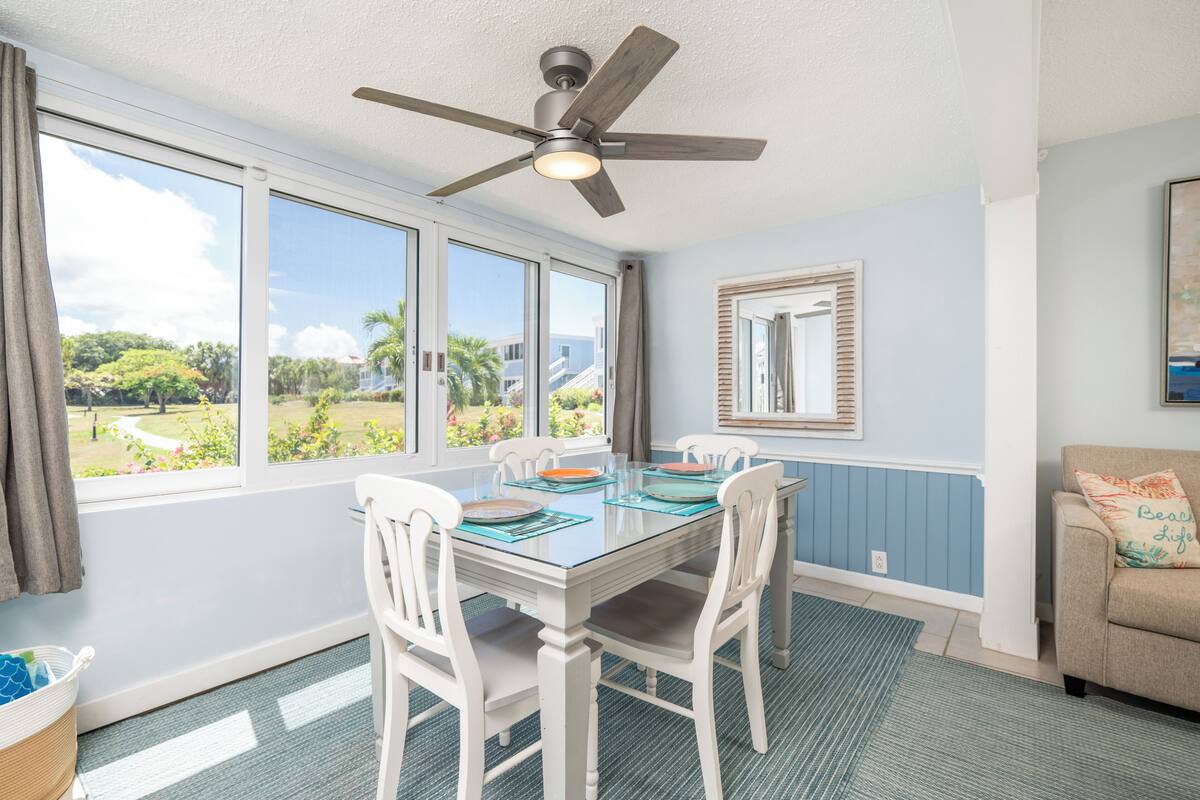 A dining area with a glass table, white chairs, a ceiling fan, and large windows with a view of greenery.
