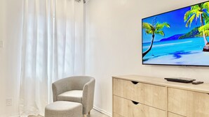 A modern living room with a flat-screen TV on the wall, a grey armchair, a small ottoman, and a wooden cabinet. - Crystal Cove Beachfront Pool Tennis (St. Thomas)