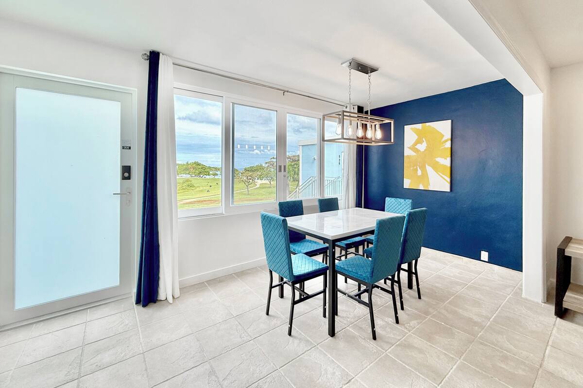 A dining area with a white table and blue chairs, large windows with a view of the sea, and a blue accent wall with a yellow abstract painting.