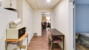 Una cocina compacta con microondas, un refrigerador pequeño y una mesa de madera con una silla. Un pasillo conduce a una sala de estar con sofá y una lámpara.