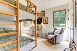 Een kamer met een stapelbed, een grijze fauteuil, een houten dressoir en ingekaderde foto's aan de muur.
