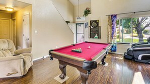 Une table de billard dans une pièce où se trouvent une chaise, un jeu de fléchettes et une fenêtre avec des rideaux.