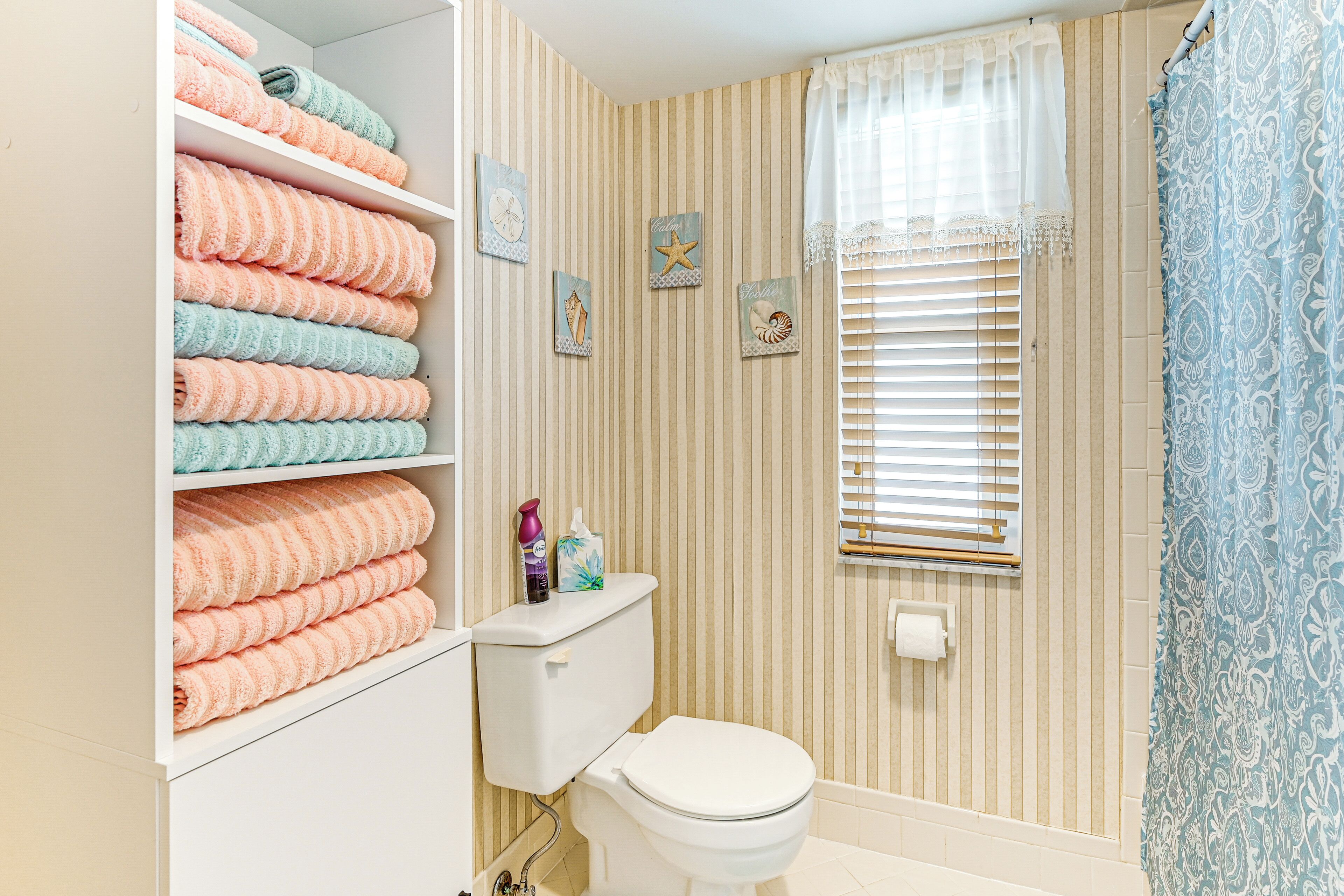 A bathroom with striped wallpaper, a toilet, a shelf with folded towels, and a window with blinds.