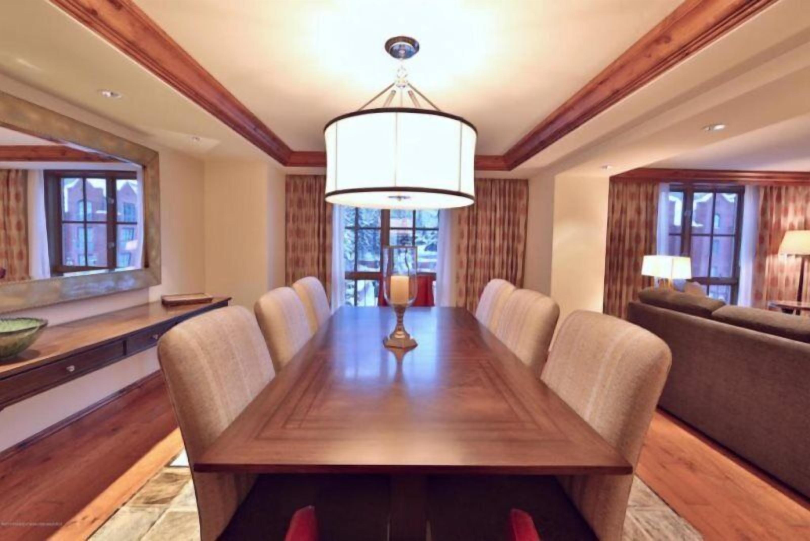 A dining room with a long wooden table, beige upholstered chairs, and a large chandelier.