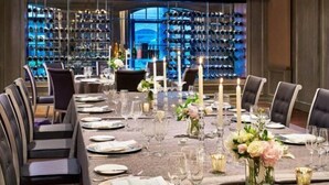 A formal dining room with a long table set for a meal, surrounded by chairs, and a wine cellar in the background.