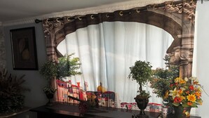 A room with a table, two potted plants, a decorative arch, and a view of a cityscape through a curtain.