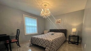 A bedroom with a bed, a desk, a chair, a nightstand, a chandelier, and a window with blinds.