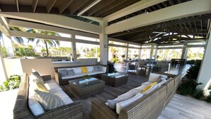An outdoor lounge area with wicker furniture and glass-top tables, surrounded by a covered patio with a view of palm trees. - Quaint villa Beach access Elina (Sabana)