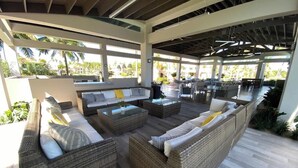 Un espace extérieur aménagé, abrité et doté de mobilier en osier et de tables à plateaux en verre, entouré d’une structure blanche avec de grandes fenêtres.