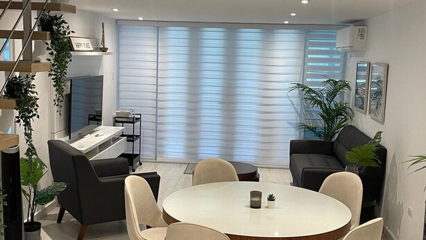 Un salon moderne avec une table à manger ronde, des sièges confortables, un escalier et une grande fenêtre équipée de stores.