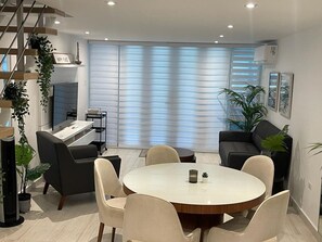 Un salon moderne avec une table à manger ronde, des sièges confortables, un escalier et une grande fenêtre équipée de stores.