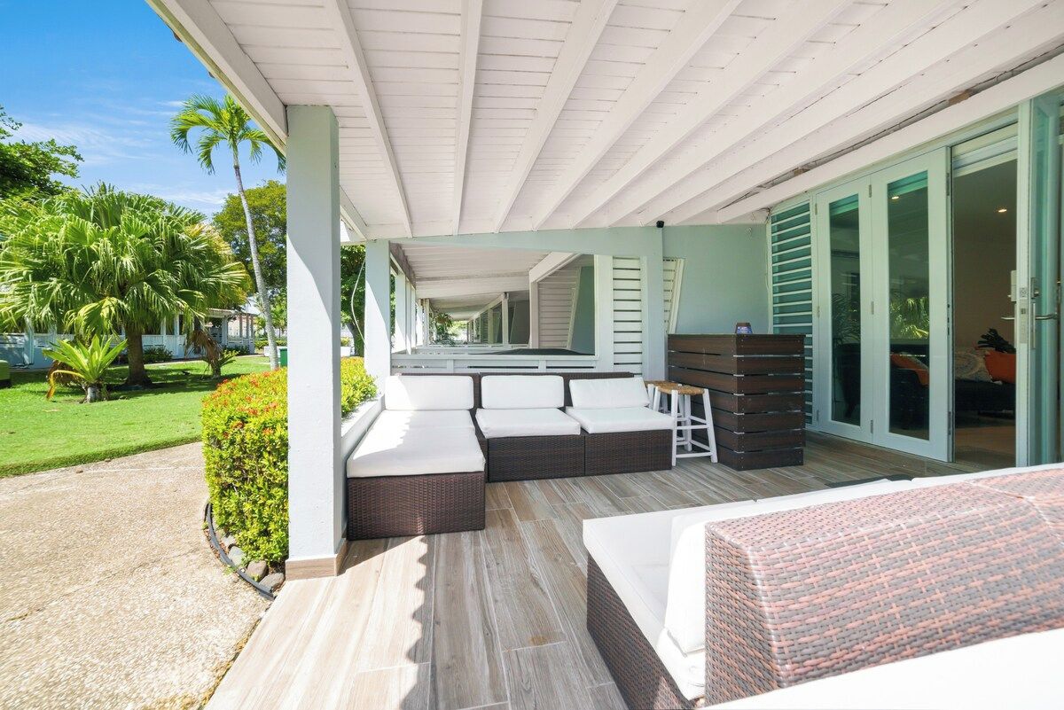 A covered outdoor seating area with a sofa and a table, surrounded by a garden with palm trees.