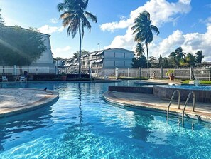 A swimming pool with a white fence, palm trees, and a building in the background.