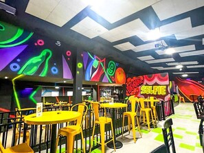 A brightly lit indoor space with yellow tables and chairs, a large colorful mural on the wall, and a ceiling with geometric patterns.