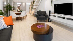 Una sala moderna con televisor de pantalla plana, comedor con sillas, sofá con cojines y escalera decorada con plantas.