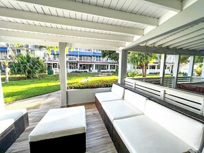 Un espace extérieur aménagé, abrité et doté de bancs blancs rembourrés et d’un plancher en bois, offrant une vue sur un jardin bien entretenu et sur des bâtiments situés au loin.