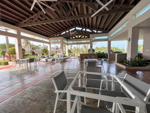 Een overdekt terras buiten met zitplaatsen en tafels, omgeven door planten en uitzicht op palmbomen.