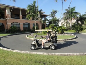 A golf cart on a paved road with a landscaped area and a building with columns in the background.
