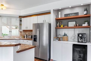 A modern kitchen with white cabinets, a stainless steel refrigerator, a built-in oven, a coffee machine, and a shelf with decorative items.