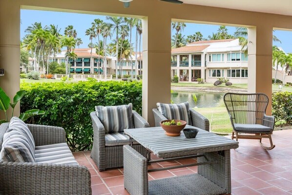 A covered outdoor seating area with wicker furniture, a table, and a view of a resort building and palm trees.