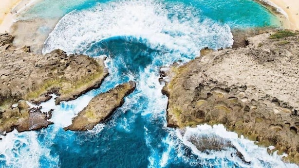 Vista aérea de una zona costera con playa, aguas azules cristalinas y formaciones rocosas.