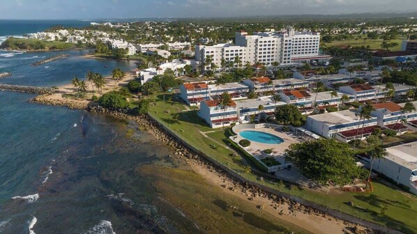 Vista aérea de un complejo turístico costero con varios edificios, una piscina y una zona de playa.