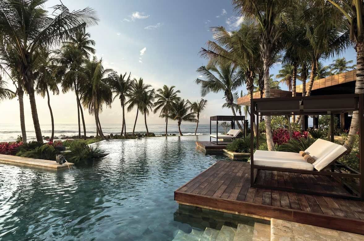 An infinity pool with a wooden deck, lounge chairs, and palm trees.