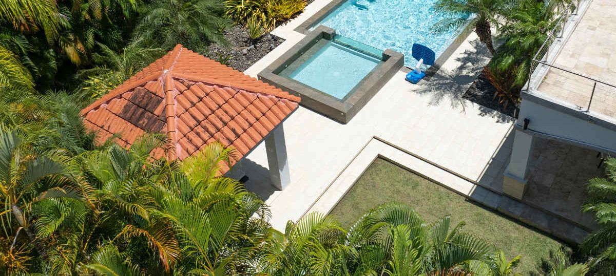 A swimming pool with a tiled deck, surrounded by palm trees and a red-roofed structure.