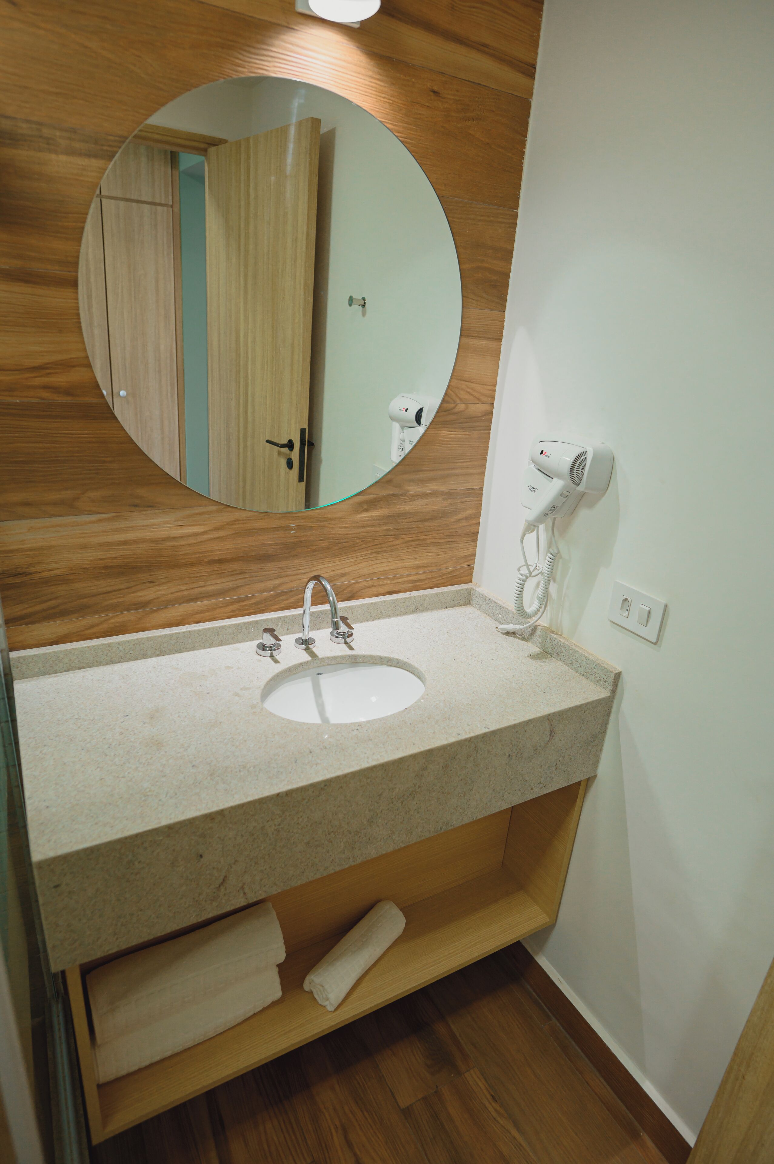 A bathroom with a wooden vanity, a round mirror, a white sink, and a hairdryer on the wall.