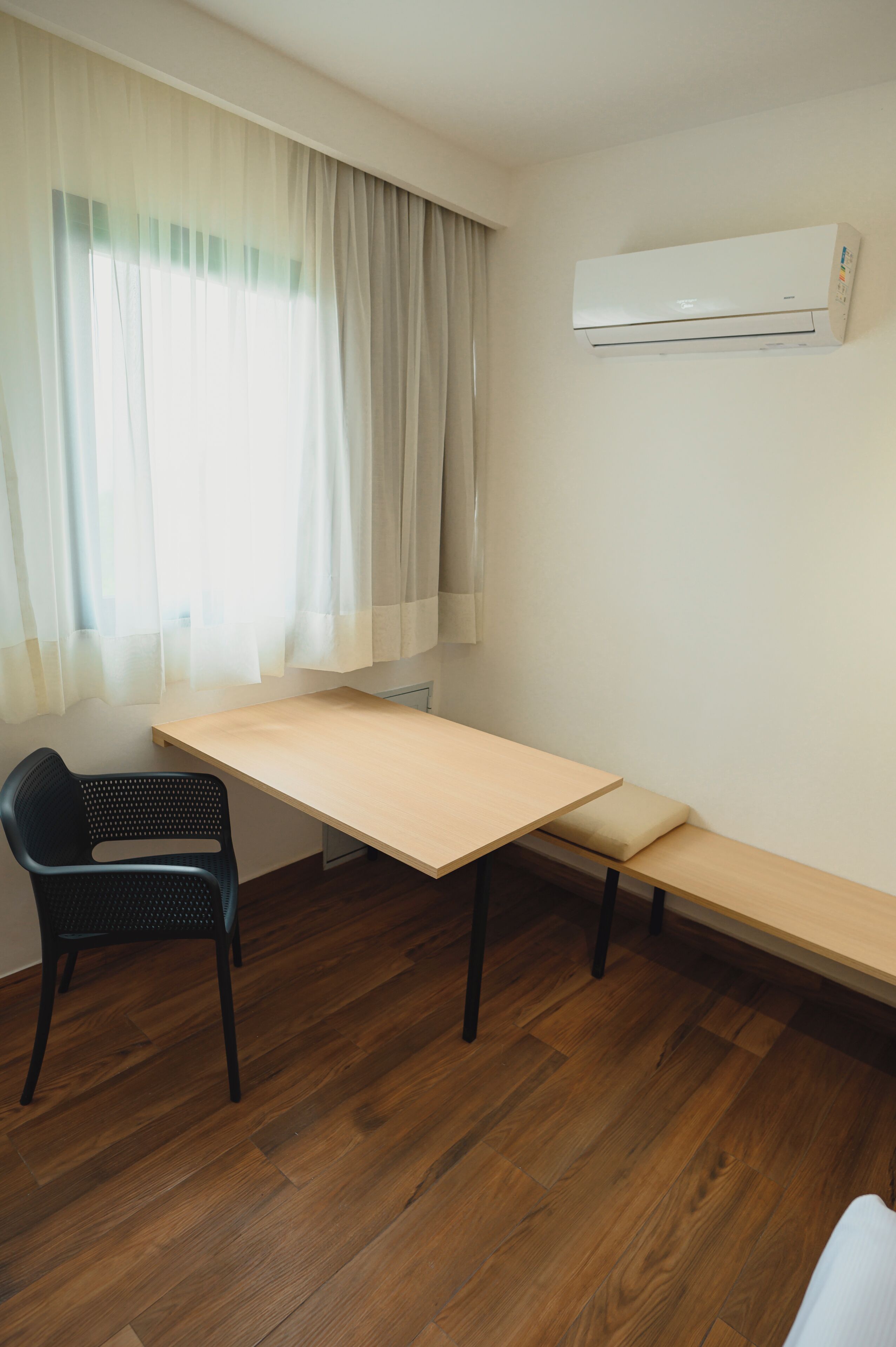 Une pièce avec un plancher en bois, un bureau avec une banquette, une chaise noire et un appareil de climatisation fixé au mur.