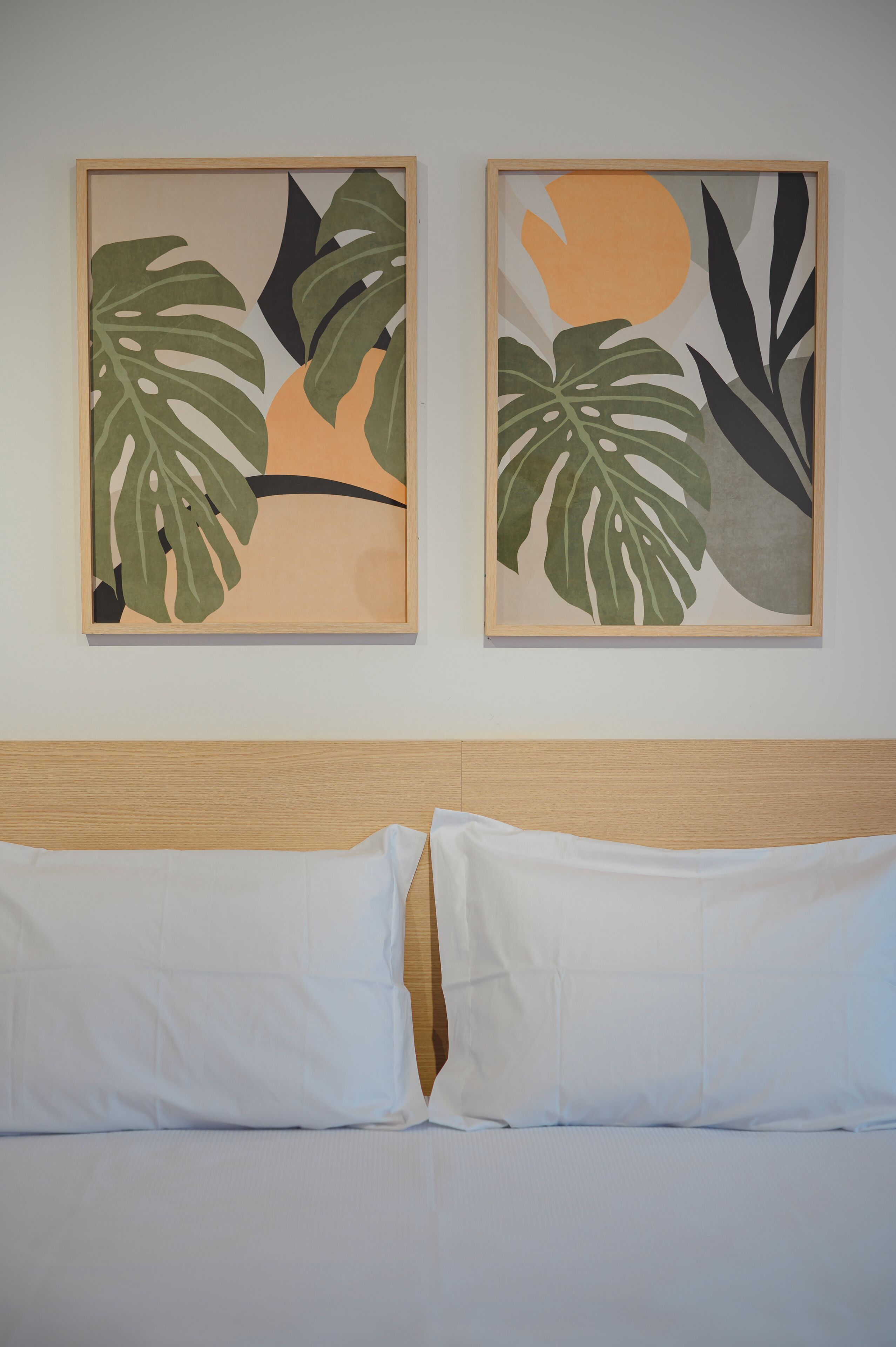 Cama con ropa blanca y cabecera de madera, flanqueada por dos cuadros con hojas tropicales y formas abstractas.