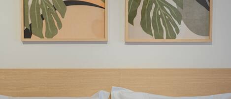 A bed with white bedding and a wooden headboard, flanked by two framed artworks depicting tropical leaves and abstract shapes.