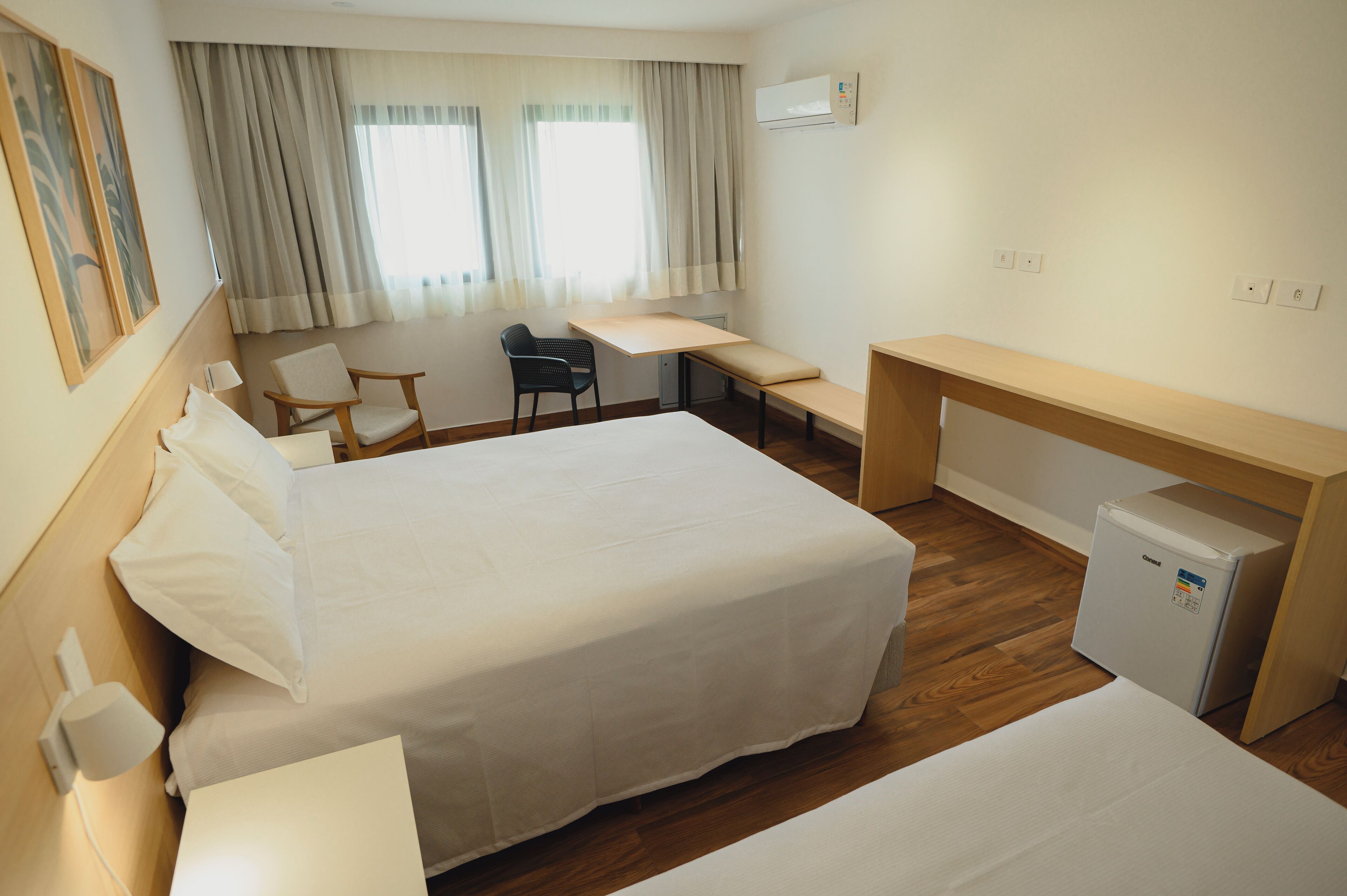 A hotel room with two beds, a desk, a chair, a small refrigerator, and a window with curtains.