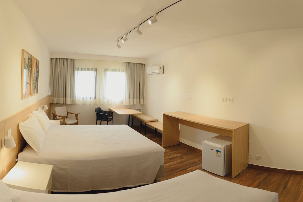 Ein Hotelzimmer mit zwei Betten, einem Schreibtisch, einem Stuhl, einem kleinen Kühlschrank und einem Fenster mit Vorhängen.