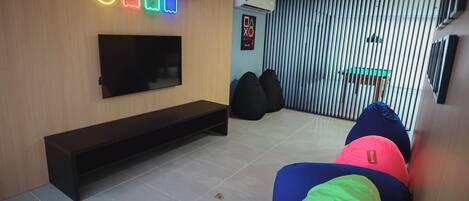 A modern living room with a flat-screen TV, a bench, bean bags, and a wall-mounted neon sign.