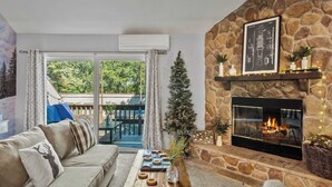A cozy living room with a stone fireplace, a sofa, a coffee table with decorative items, and a view of the outdoors through a glass door. - Slopeside Escape by Avantstay Walk to Ski Lifts Arcade Games + Shuffleboard (Tannersville)