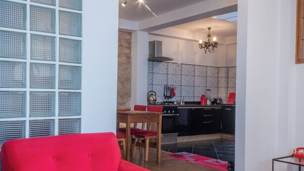 A modern living room with a red sofa, a dining area with a wooden table and chairs, and a kitchen with black cabinets and a tiled backsplash.
