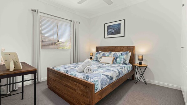 A bedroom with a wooden bed, a desk with a lamp, and a window with blinds.