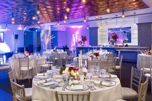 Uma sala de jantar bem iluminada com mesas redondas preparadas para um evento formal, com toalhas brancas, talheres e arranjos florais.