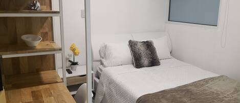 A bedroom with a bed, a bedside table, a shelf with a bowl and a plant, and a window.