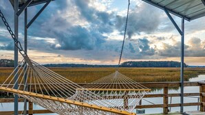 A hammock on a wooden deck overlooking a marshy landscape with a cloudy sky. - The Dock House (Savannah)