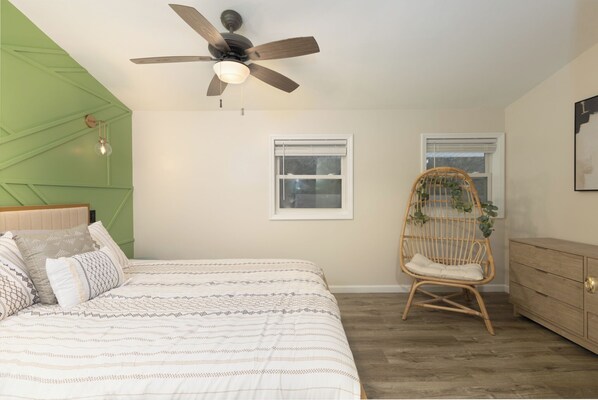 A bedroom with a bed, a ceiling fan, two windows, a wicker chair, and a wooden dresser.