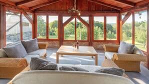 A spacious room with a high ceiling and large windows offering a view of a forested landscape. There are wicker sofas and a central table with glasses and a bottle.