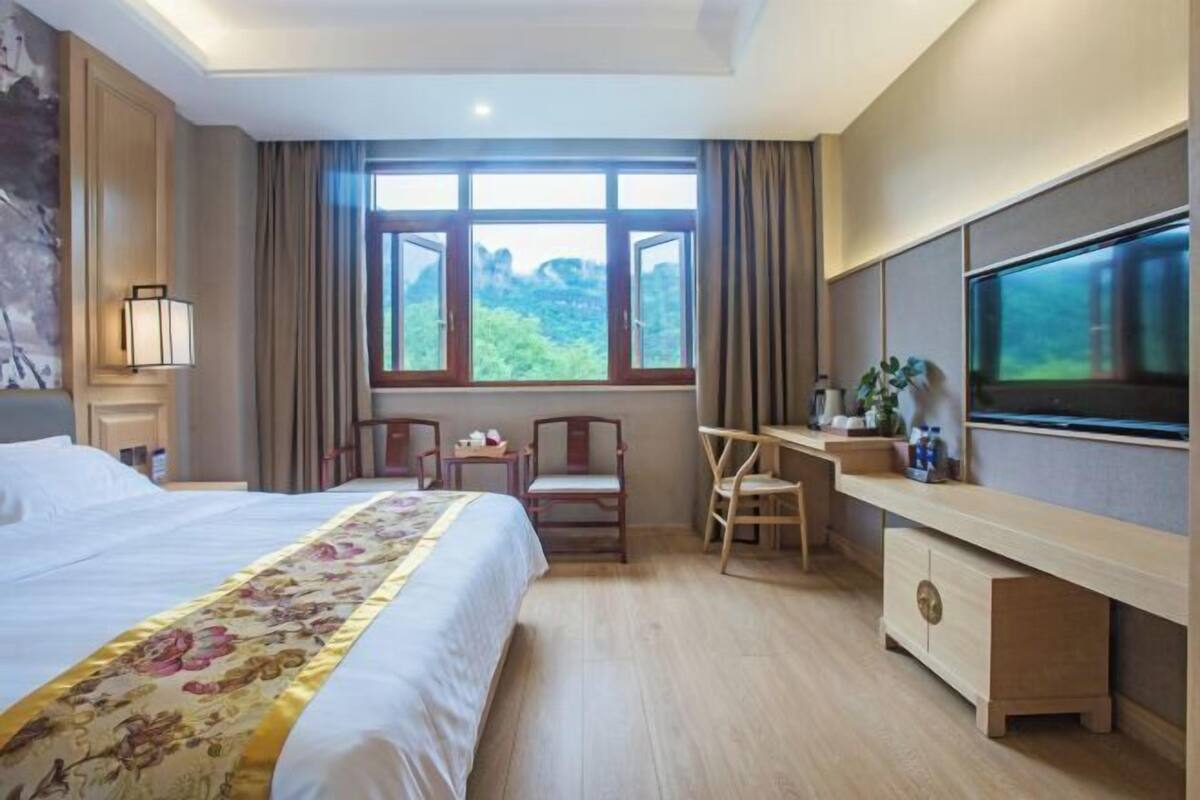 A hotel room with a bed, a desk, a TV, and a window with a view of greenery.