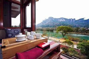 A wooden table with a red cushion, white teapots, and a teapot set on a window overlooking a river and mountains. - Wuyishan Wuyi Yard Chinese Inn (Wuyishan)